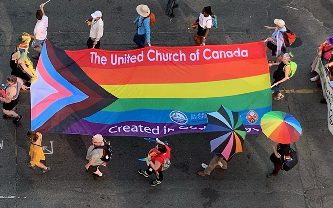 Rainbow flag with the words Created in the image of God carried by 12 people in the Pride parade